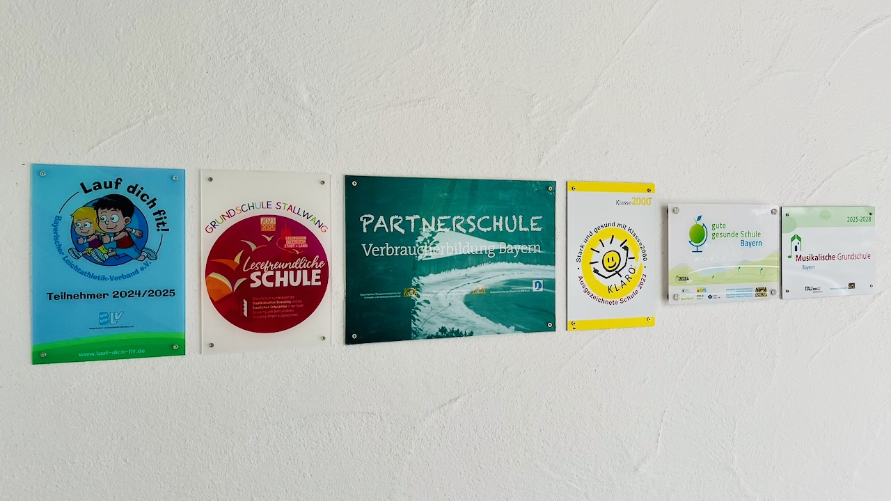 Verschiedene Logotafeln an einer weißen Wand: Musikalische Grundschule. Lesefreundliche Schule, TfK, Lauf dich fit, Klara